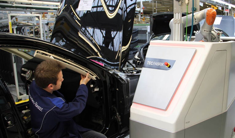 Mitarbeiter arbeitet gemeinsam mit einem kollaborativen Roboter und einer industriellen Maschinenverkleidung in der Fahrzeugmontage bei Mercedes-Benz.